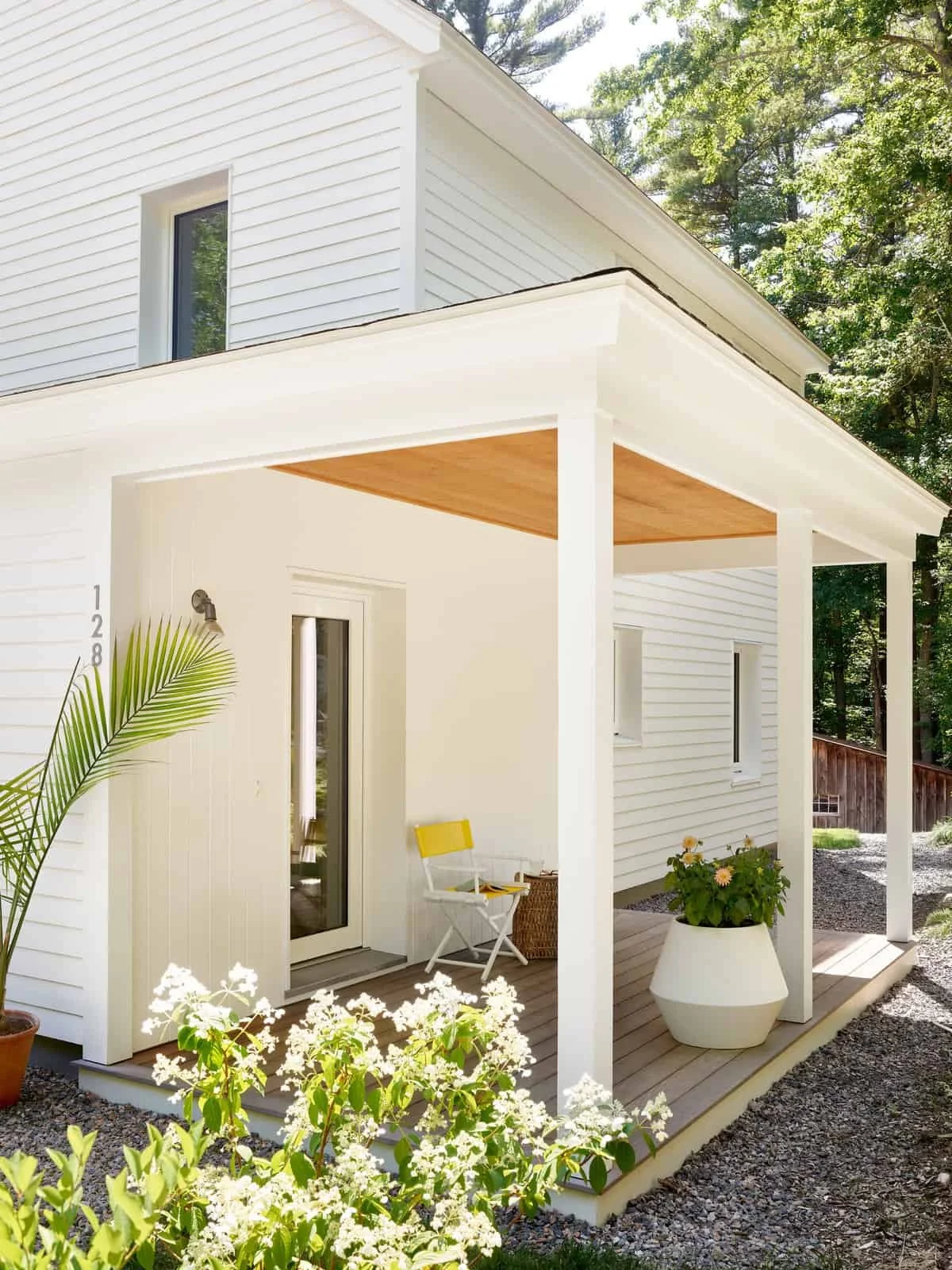 A modern white house with a small covered porch area, decorated with potted plants and outdoor furniture, surrounded by lush green trees.