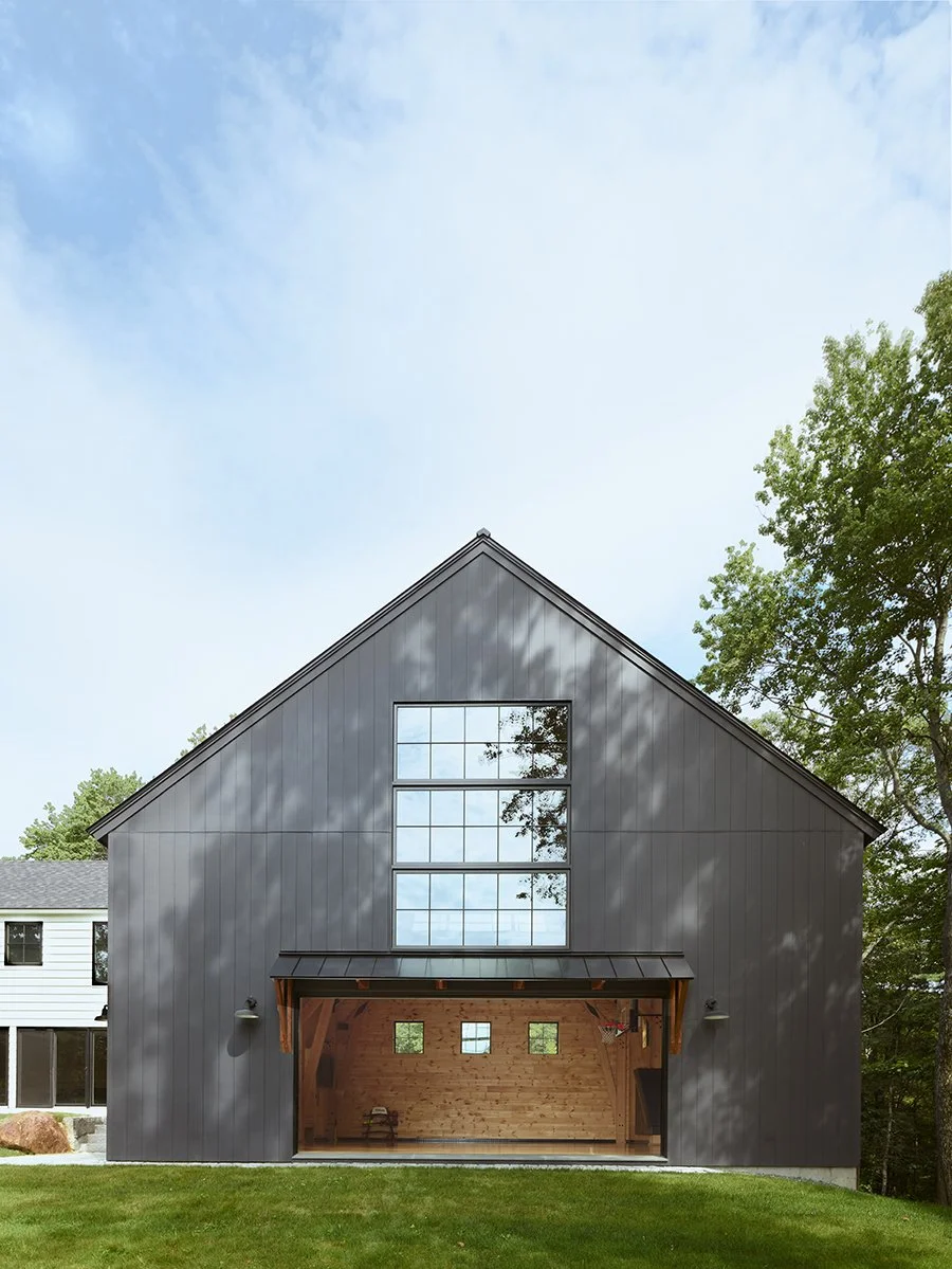Modern barn-style house with black metal exterior and large windows, with open garage showcasing wooden beam interior, surrounded by grass and trees under a partly cloudy sky.