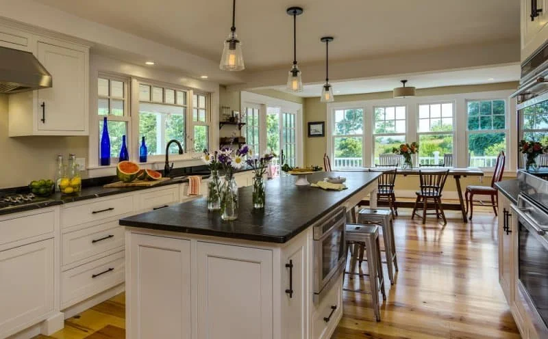 Bright, spacious kitchen with white cabinets, black countertops, and large windows overlooking a green outdoor area. The central island features flowers in glass jars and kitchenware, with pendant lights hanging above. There is a dining area with woo