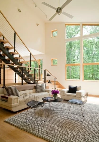 Living room with large windows showing green trees, a beige sofa with striped cushions, two white armchairs, a black coffee table with a purple flower arrangement, and modern black wire chairs, with a staircase and ceiling fan.
