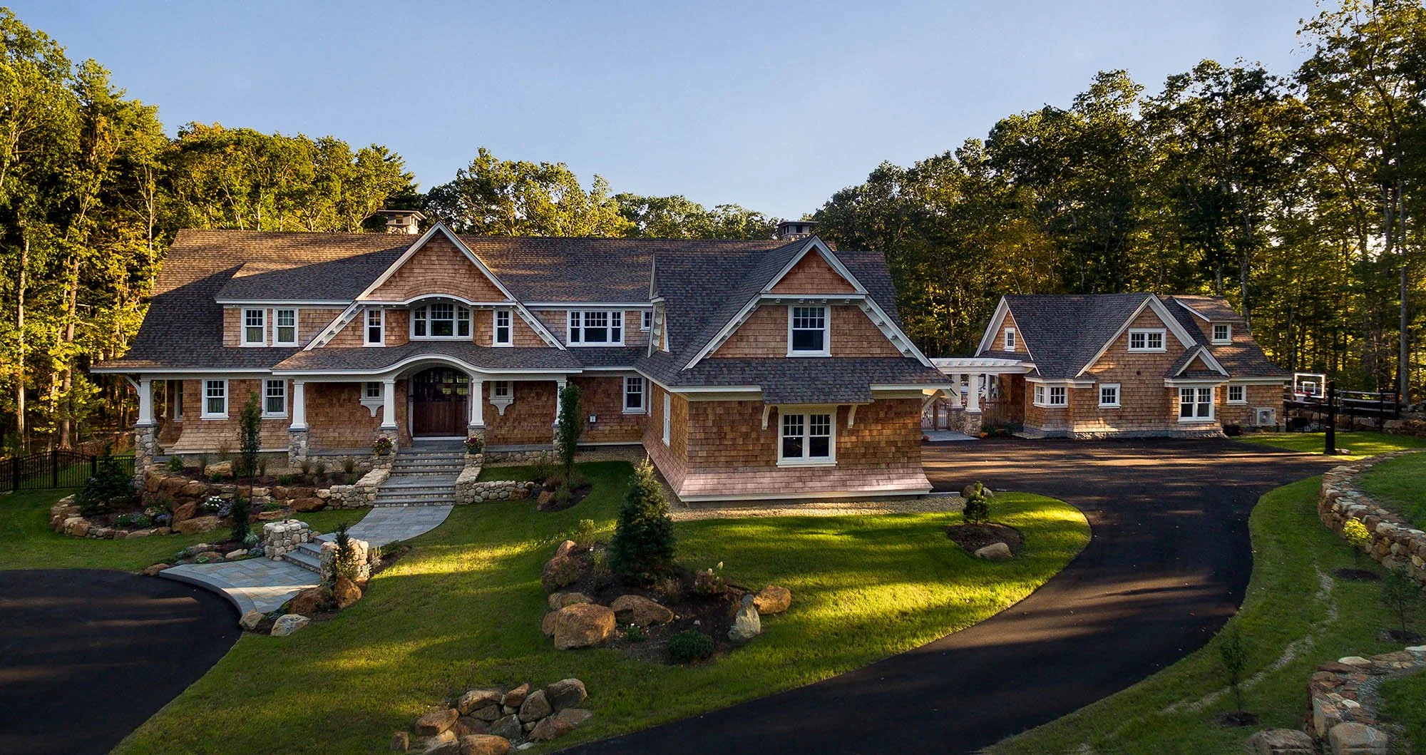Large house with a front porch, surrounded by greenery and trees, with a curved driveway, stone landscaping, and a basketball hoop in the backyard.