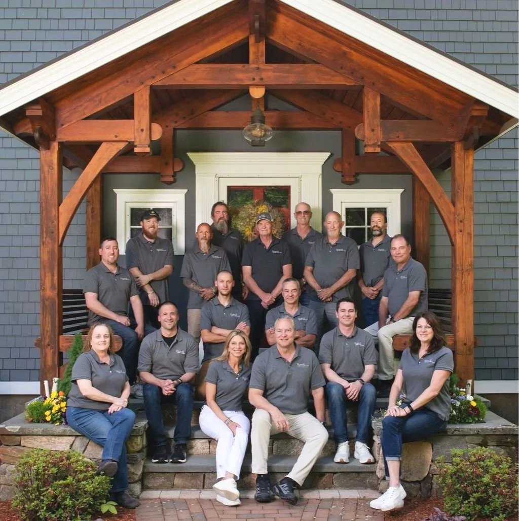 Howell Custom Building Group's team of 15 people, wearing matching gray shirts, posing on the steps of a house with a wooden porch and a painted floral wreath on the door.