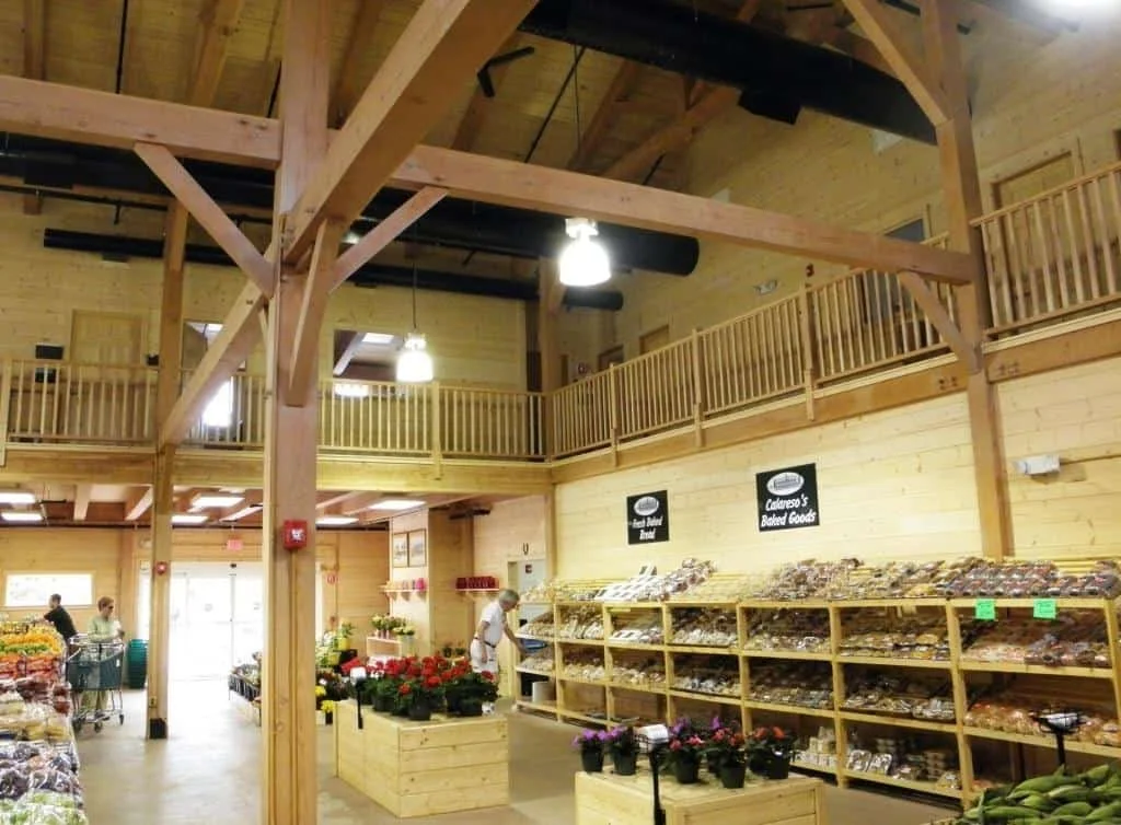 Inside a grocery store with wooden interior, shelves filled with baked goods, flowers, and various products, customers shopping, and a second level balcony.