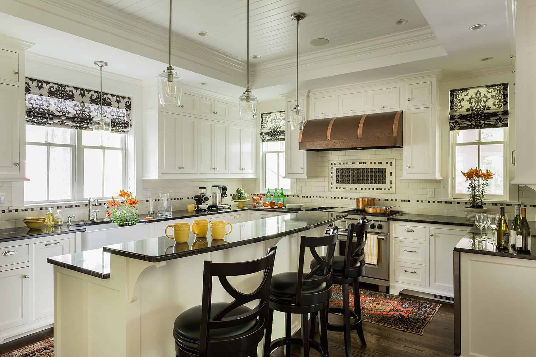 A modern kitchen with white cabinets, black countertops, and pendant lights. There are windows with black and white patterned blinds, and a kitchen island with yellow mugs and black chairs. The kitchen has plants, bottles, and cooking utensils.