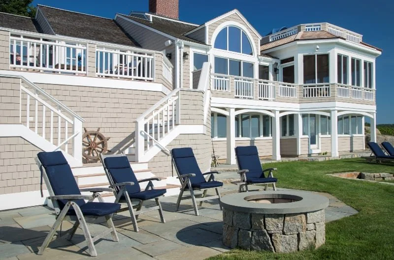 A large white house with multiple levels, large windows, and a balcony. In front are five blue lounge chairs and a round stone fire pit on a stone patio. Green lawn surrounds the patio.