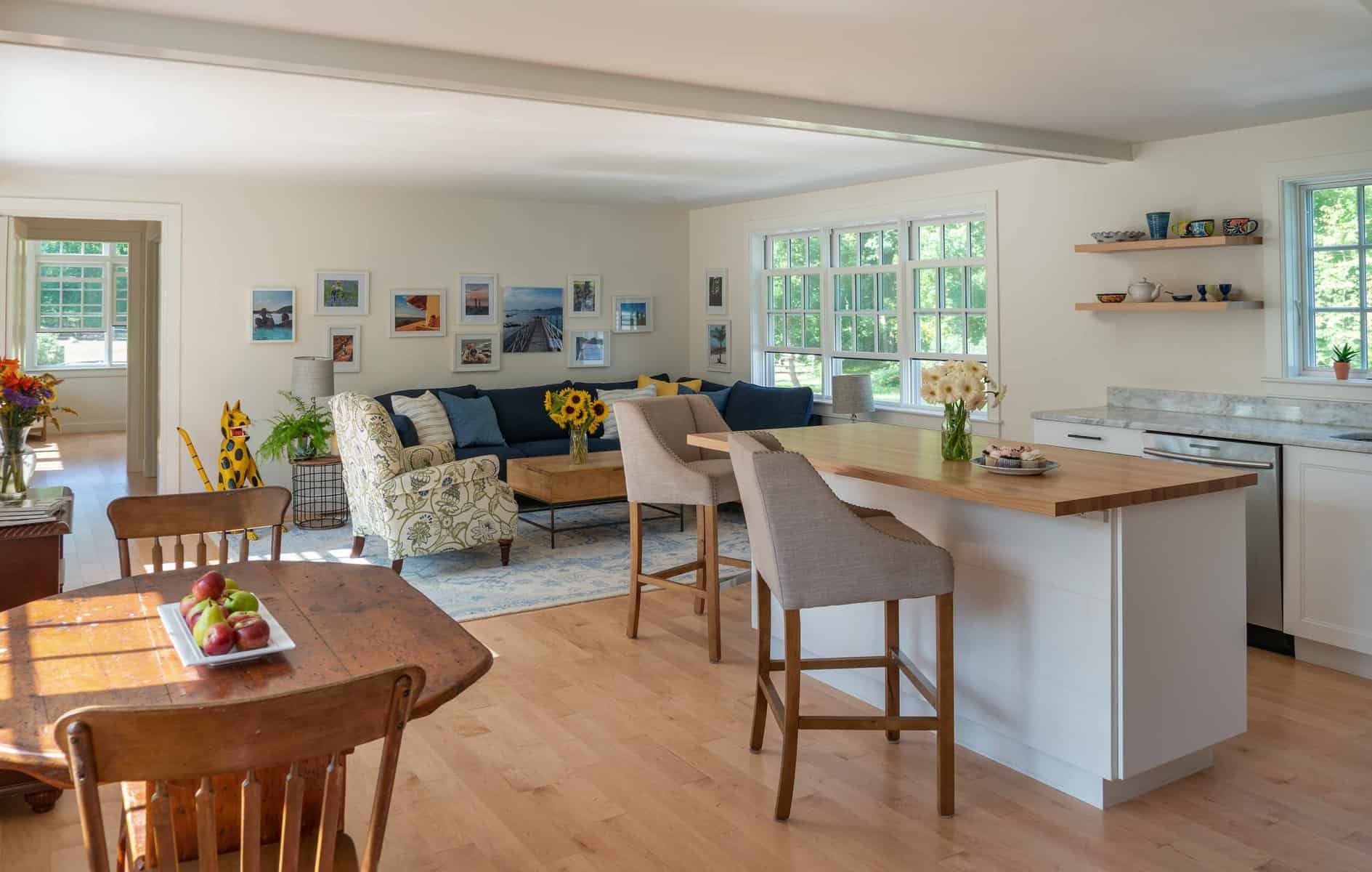 Living room with large windows, a dark blue sofa, a beige armchair, a wooden coffee table with sunflower bouquet, and a gallery wall of framed photos. Adjacent kitchen area with a wooden dining table, chairs, and a breakfast bar with flowers and cupc