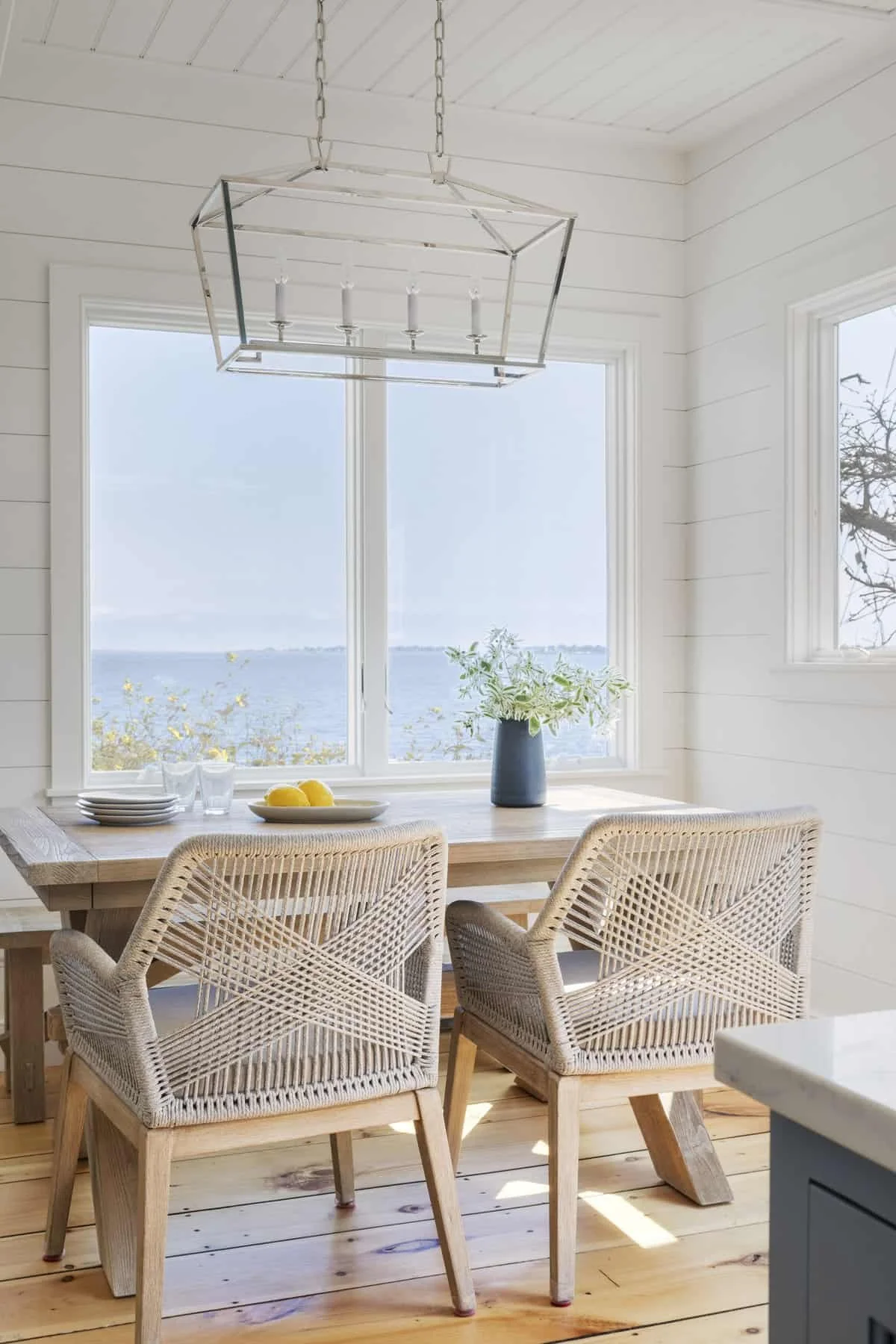 Dining area with a wooden table, four wicker chairs, a large window with a view of water, a blue vase with green plants, a white plate with lemons, empty glasses, and a modern geometric chandelier hanging from the ceiling.