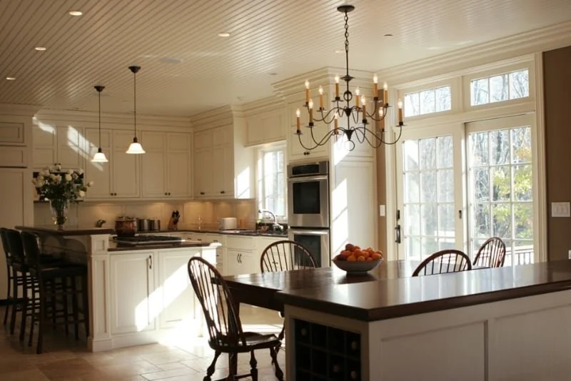 Bright kitchen with white cabinets, large windows, and a chandelier overhead. There's a kitchen island with stools, and a dining table with chairs near a sliding glass door. A bowl of oranges is on the table, and a vase with flowers is on the counter
