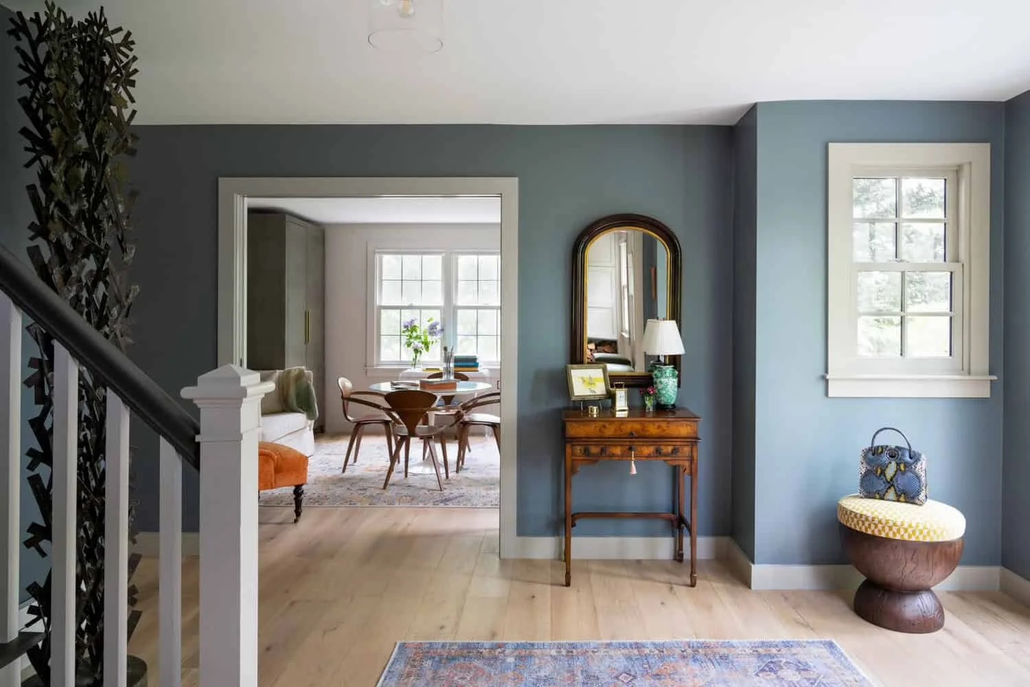 Interior view of a home's foyer with a blue wall, a wooden side table with a mirror, a table lamp, and decorative items. A window with white trim is on the right, and a round seating area with a bag on top is below it. The adjacent room shows a dinin
