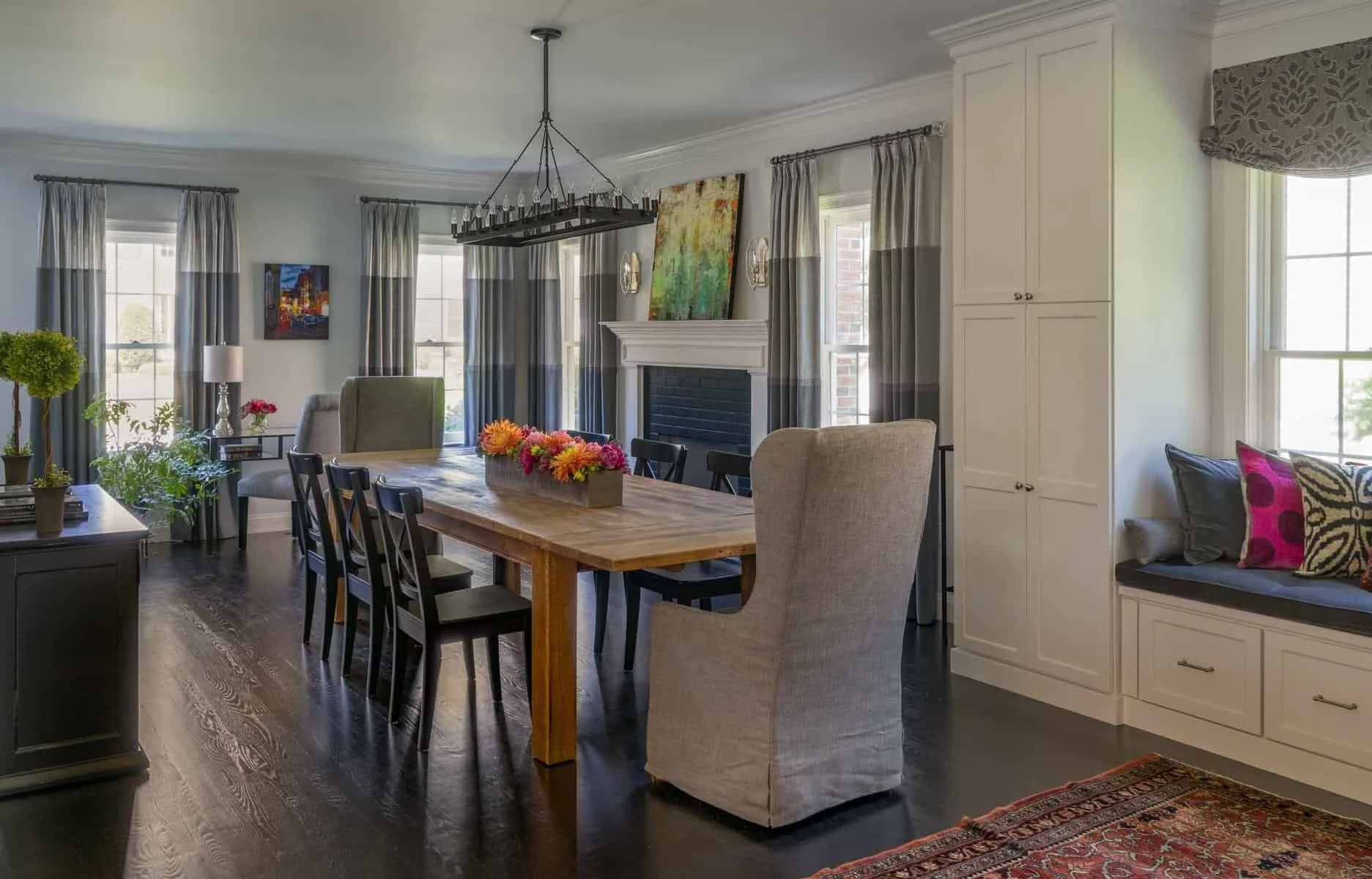 Dining room with wooden table, black chairs, beige and gray upholstered armchairs, houseplants, large windows with gray curtains, white built-in cabinets, a fireplace with a colorful painting above, chandelier, and window seat with pillows.