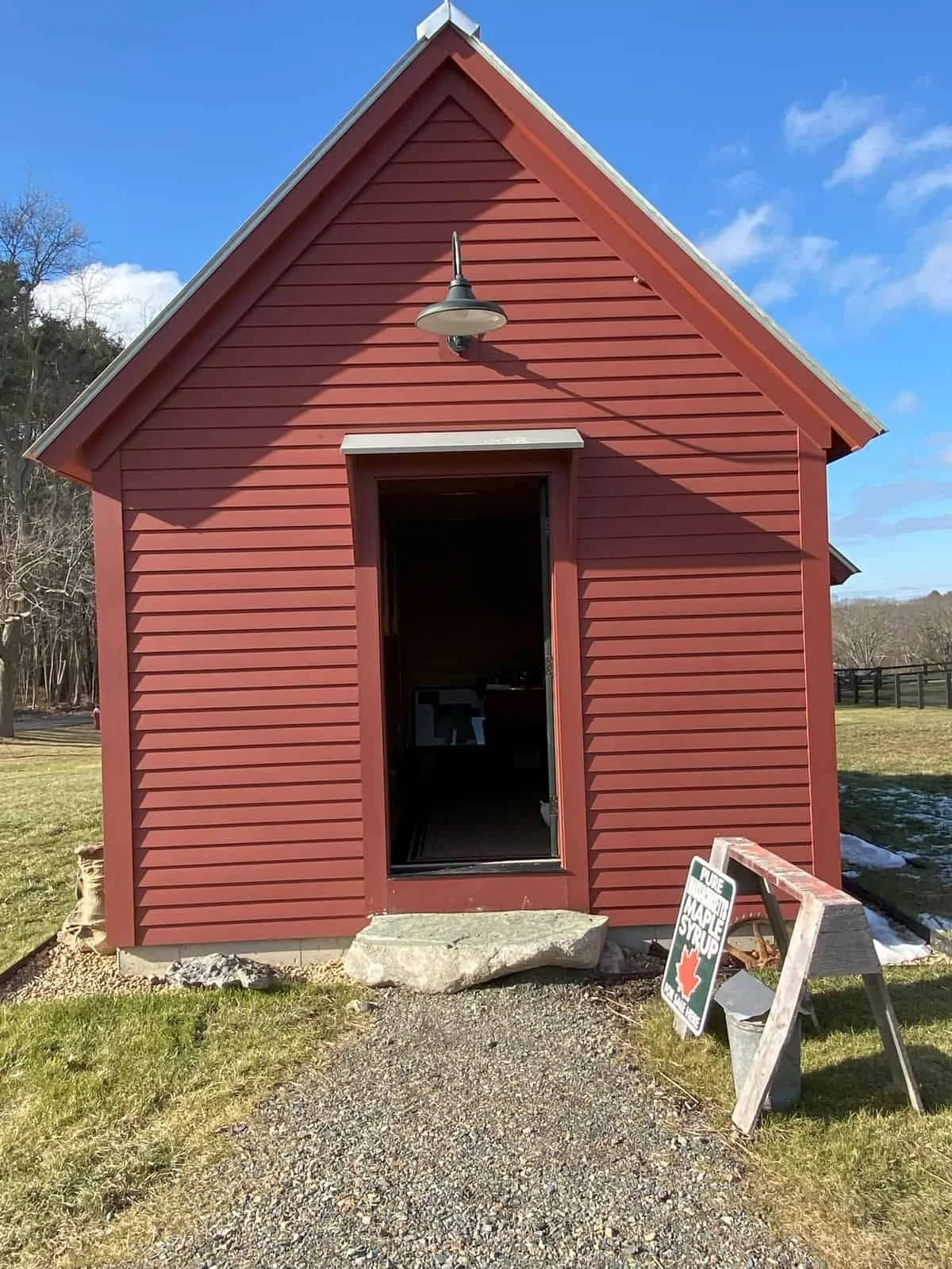 Red wooden barn with open door, black roof, and outdoor light fixture, set on a grassy area with a clear blue sky.