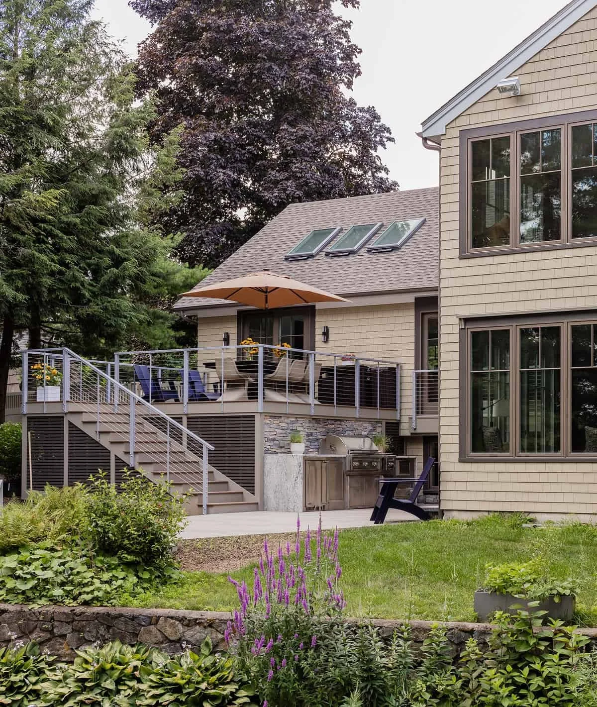 Backyard of a house with a second-story deck outdoor furniture, an umbrella, stairs, grill, and garden with purple flowers and greenery.
