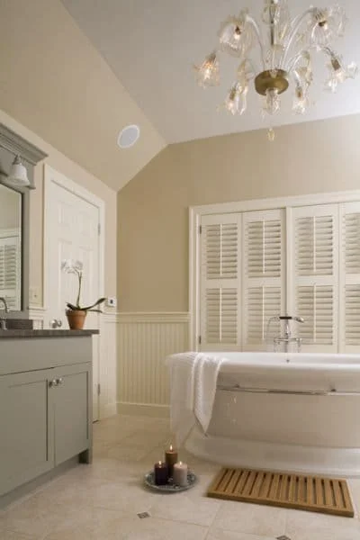 A bathroom with a freestanding bathtub, a gray vanity with a mirror, a chandelier, and a window with white shutters.