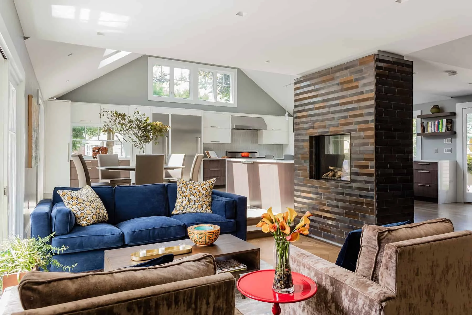 Open concept living room with blue sofa, brown armchairs, floral pillows, and a wooden coffee table with candles. A tall glass vase with orange flowers is on a small red side table. In the background, there's a dining area with a round table, four be