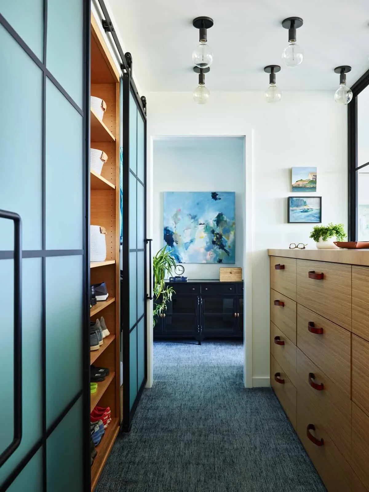 View of a modern, brightly lit room with artwork on the walls, a black console table with plants, a wooden dresser, and a sliding closet with frosted glass doors.