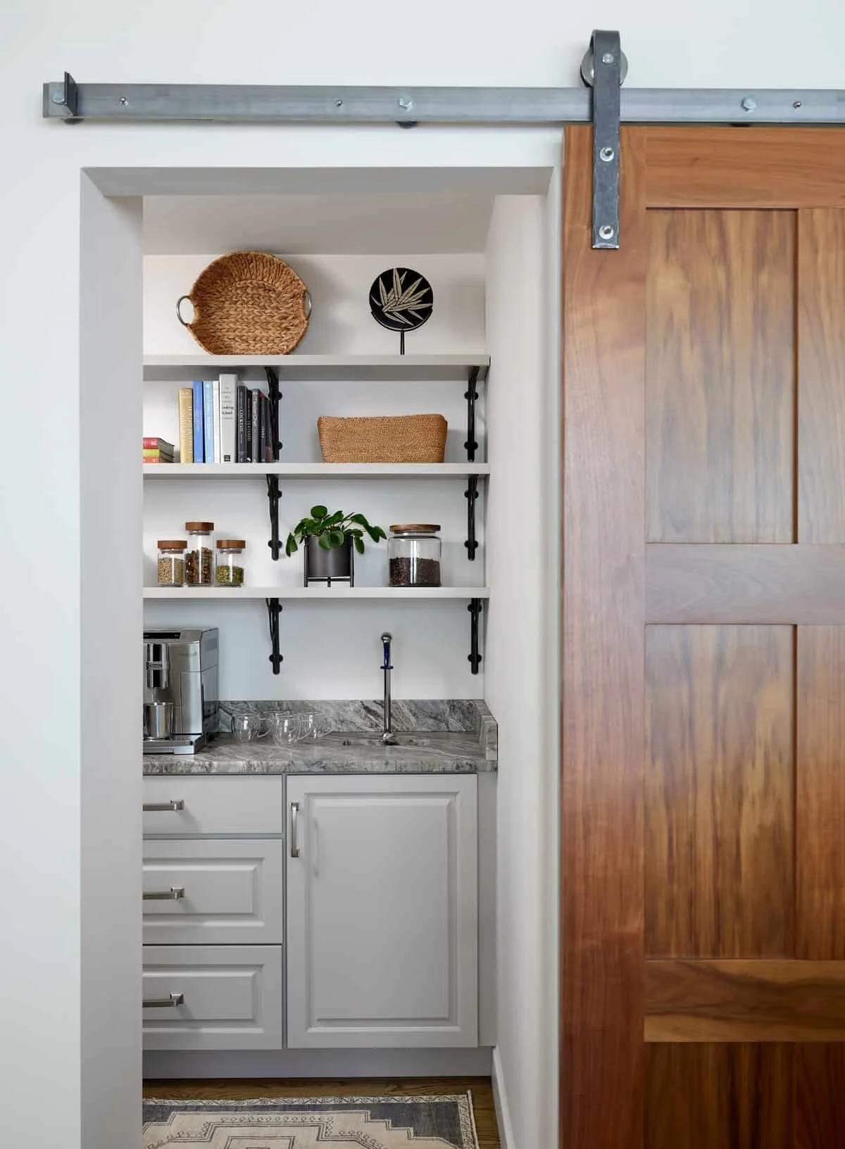 Interior view of a kitchen nook with a sliding barn door, white cabinetry with a marble countertop, open shelves with decorative items and jars, and a coffee machine.