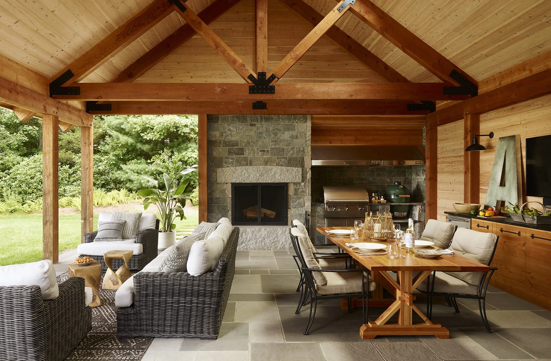 Open-concept outdoor living space with wooden ceiling and beams, featuring a stone fireplace, a seating area with wicker and cushioned chairs, and a dining area with a wooden table and cushioned chairs, surrounded by green outdoor landscape.