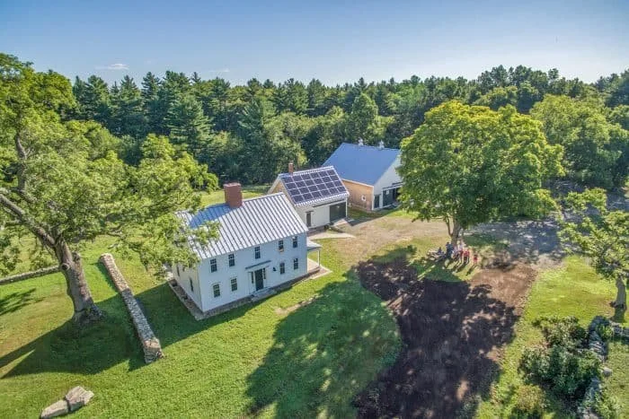 Aerial view of a rural property with a white house featuring solar panels, a large barn, and a grassy yard surrounded by trees.