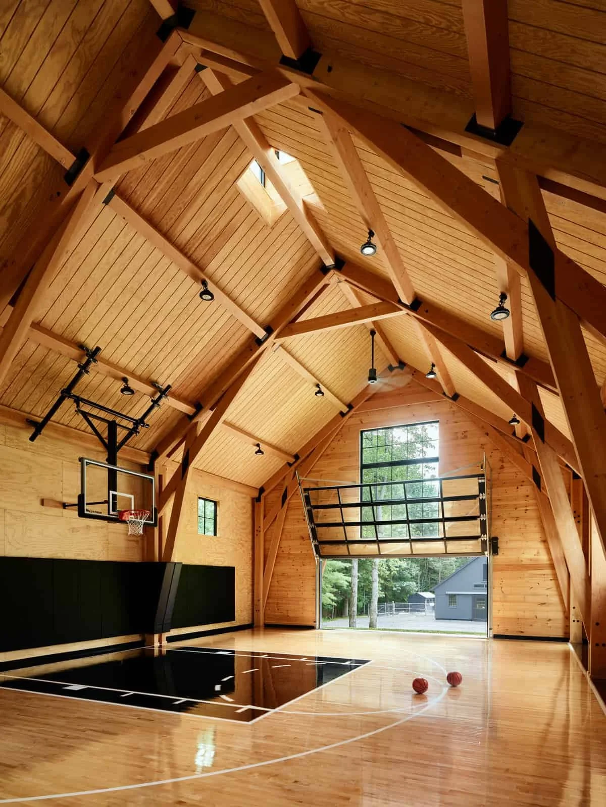 The interior of a wooden barn that functions as a gymnasium with basketball hoop and court, high ceiling with exposed wooden beams, windows letting in natural light, and two basketballs on the court.