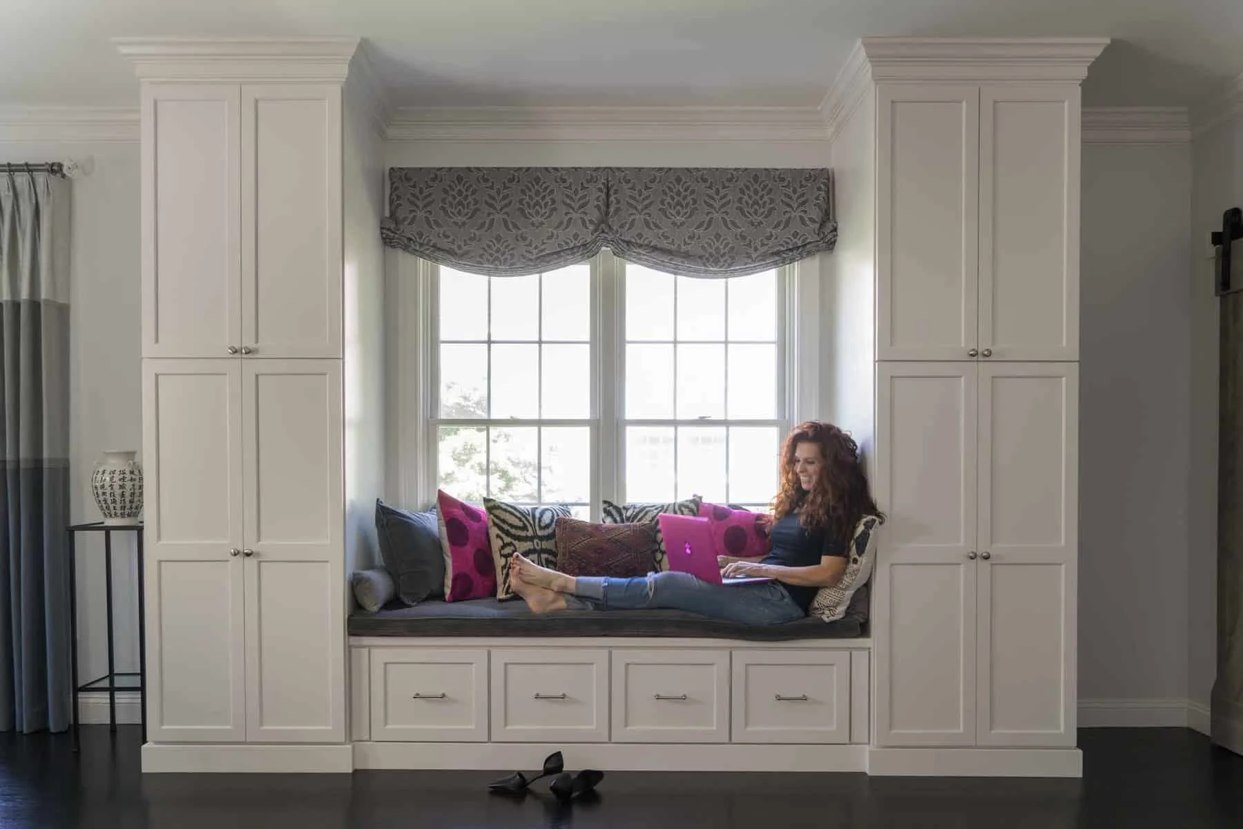 A woman with curly hair sitting on a window seat surrounded by colorful pillows, working on a laptop, in a room with built-in cabinets and a large window with valance. Shoes on the floor.
