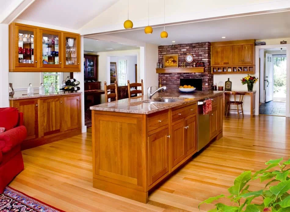 Kitchen with wooden cabinets and an island, brick fireplace, granite countertops, and hardwood floors. There's a red couch partially visible on the left and a door leading outside on the right.