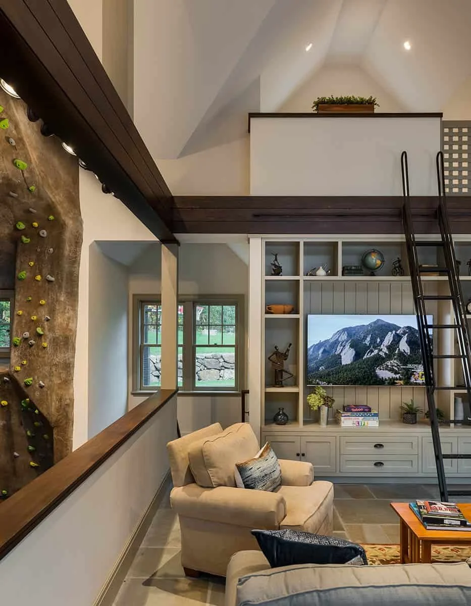 Living room with beige armchairs, a wooden coffee table with magazines, a built-in white entertainment center with books, decorative items, and a TV displaying mountains. A window shows an outdoor stone wall and greenery. A hanging rock climbing wall
