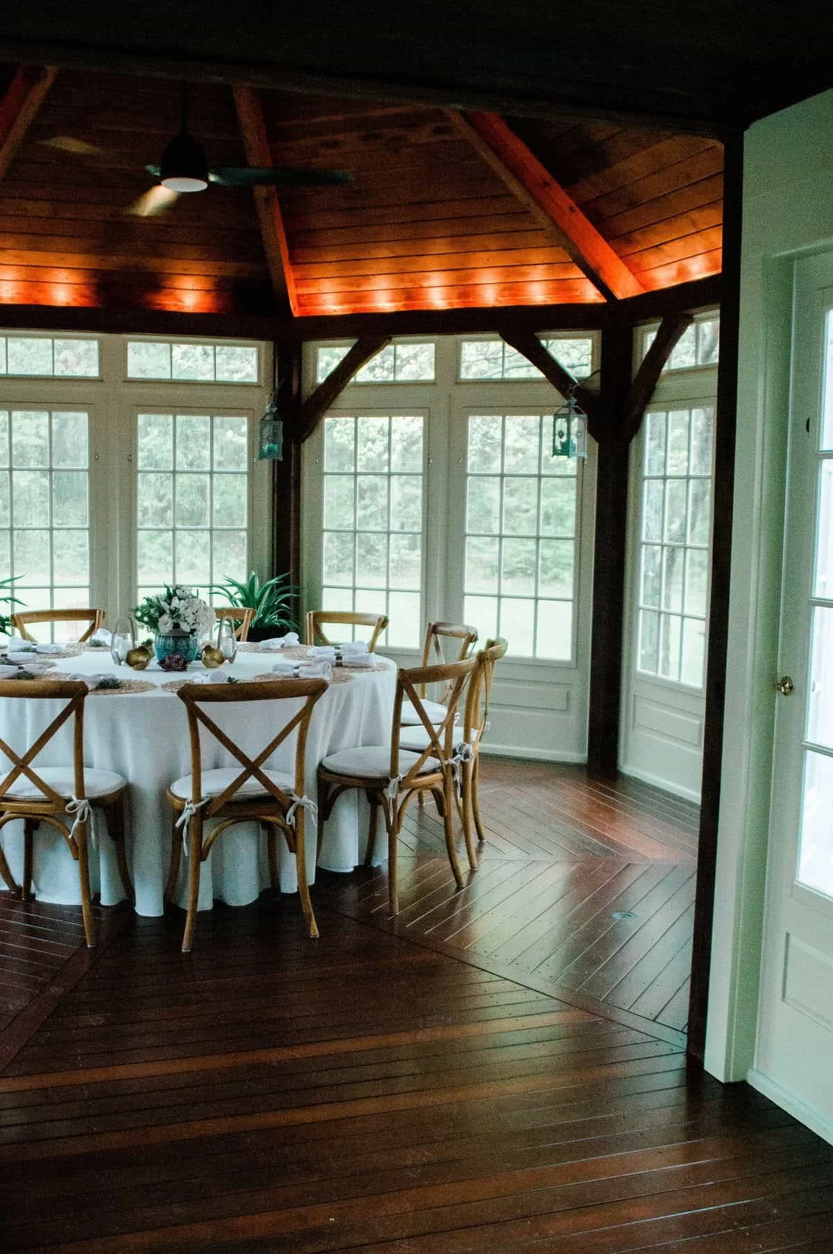 Round dining table with white tablecloth, floral centerpiece, and wooden chairs in a sunlit room with large windows and wooden ceiling beams.