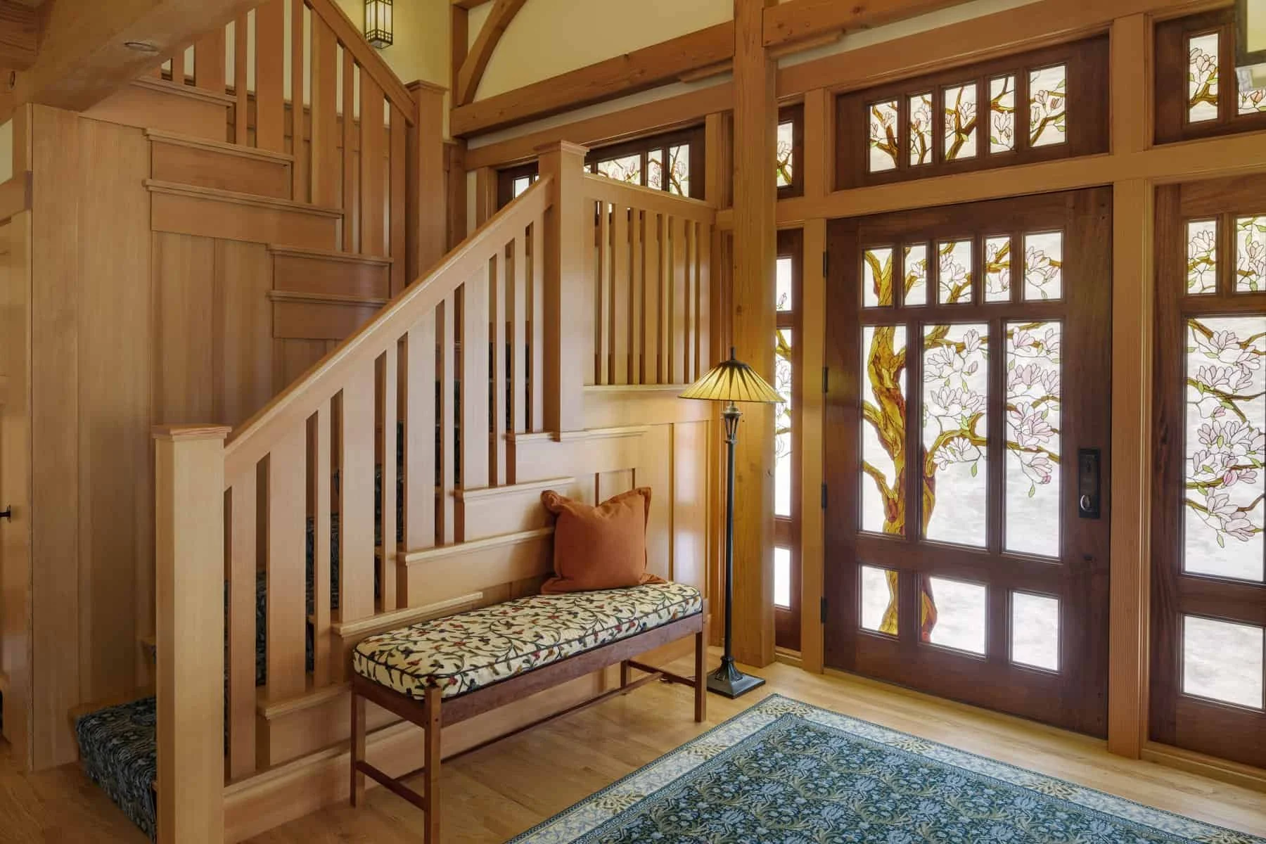 Interior view of a wooden entryway with a staircase, a window with stained glass depicting tree branches and blossoms, a bench with an embroidered cushion, a standing lamp, and a decorative rug.