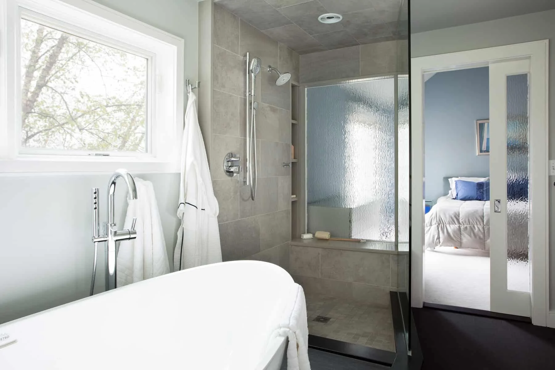 A modern bathroom with a large window, a walk-in shower with a textured glass door, and a white bathtub. Visible through a sliding door is a bedroom with a bed dressed in white and blue bedding.