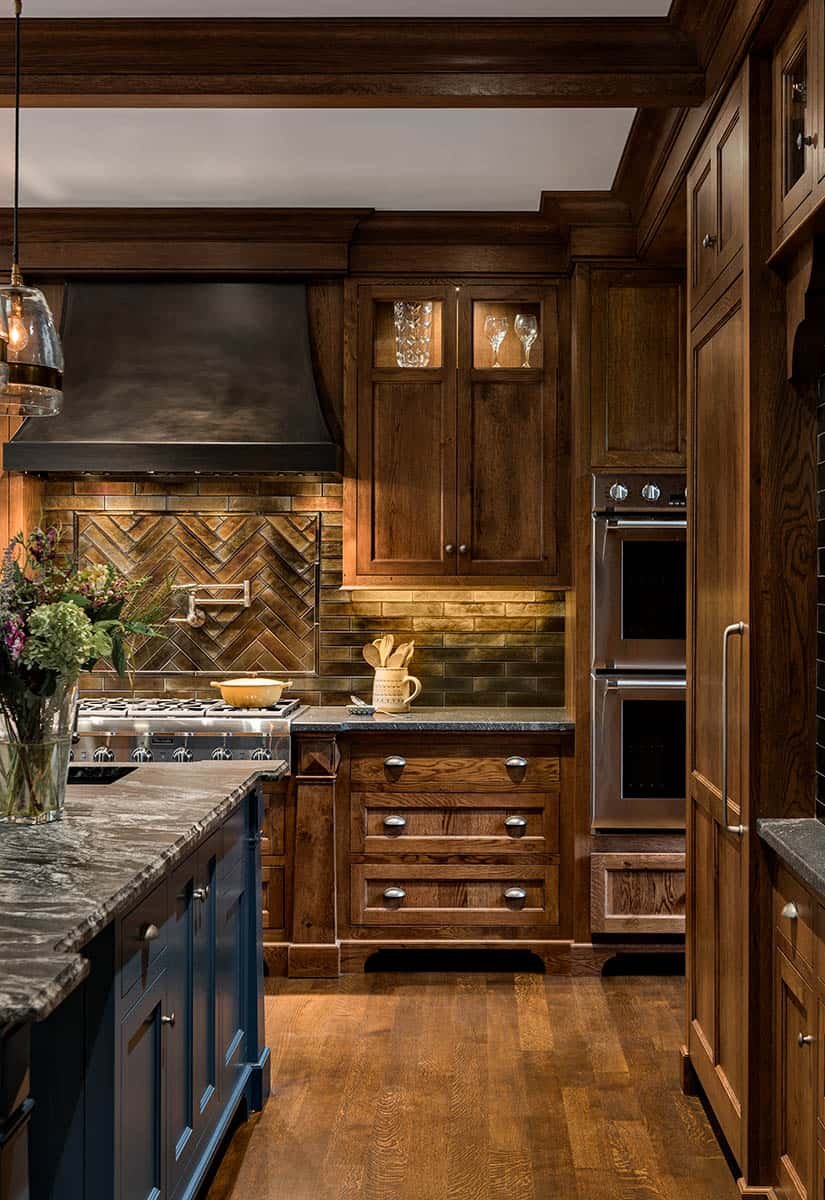 A kitchen with dark wood cabinets, a black vent hood, and a brick backsplash. There are glassware in a cabinet, a floral arrangement, and a yellow dish on the stove.