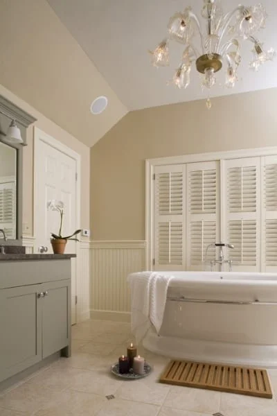 A bathroom with a freestanding bathtub, white shutters, a gray vanity, a chandelier, candles, and a potted plant.