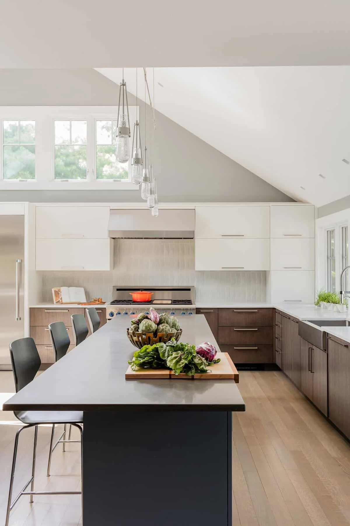 Modern kitchen with natural light, white cabinets, a large gray island, and wooden drawers. A bowl of vegetables on the island and a red pot on the stove. Large window with trees outside. Hanging glass pendant lights.