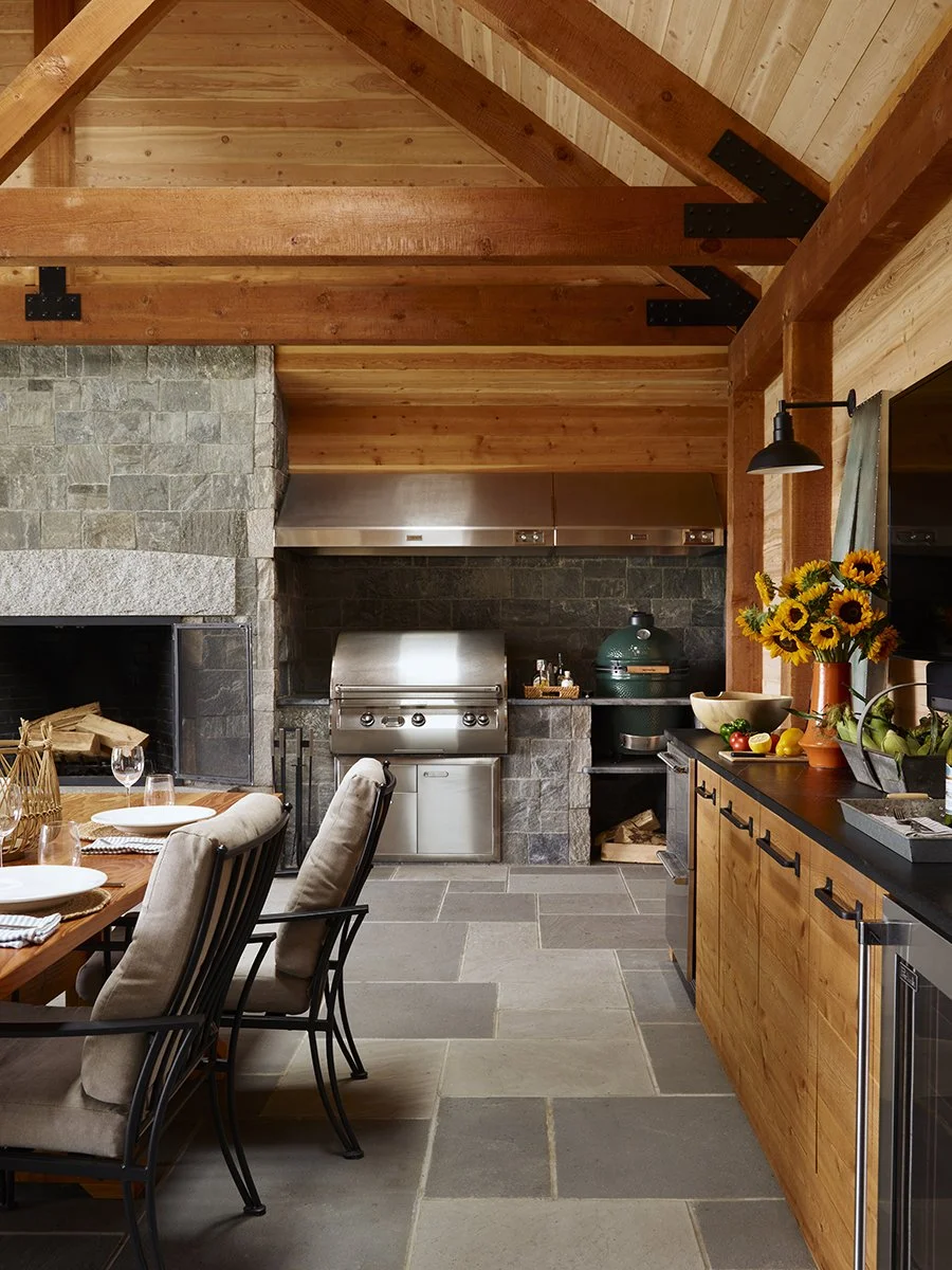 Indoor kitchen with wooden beams, a stone fireplace, a stainless steel grill, and a dining table with beige chairs. A countertop has a vase of sunflowers, bowls, and fruits.