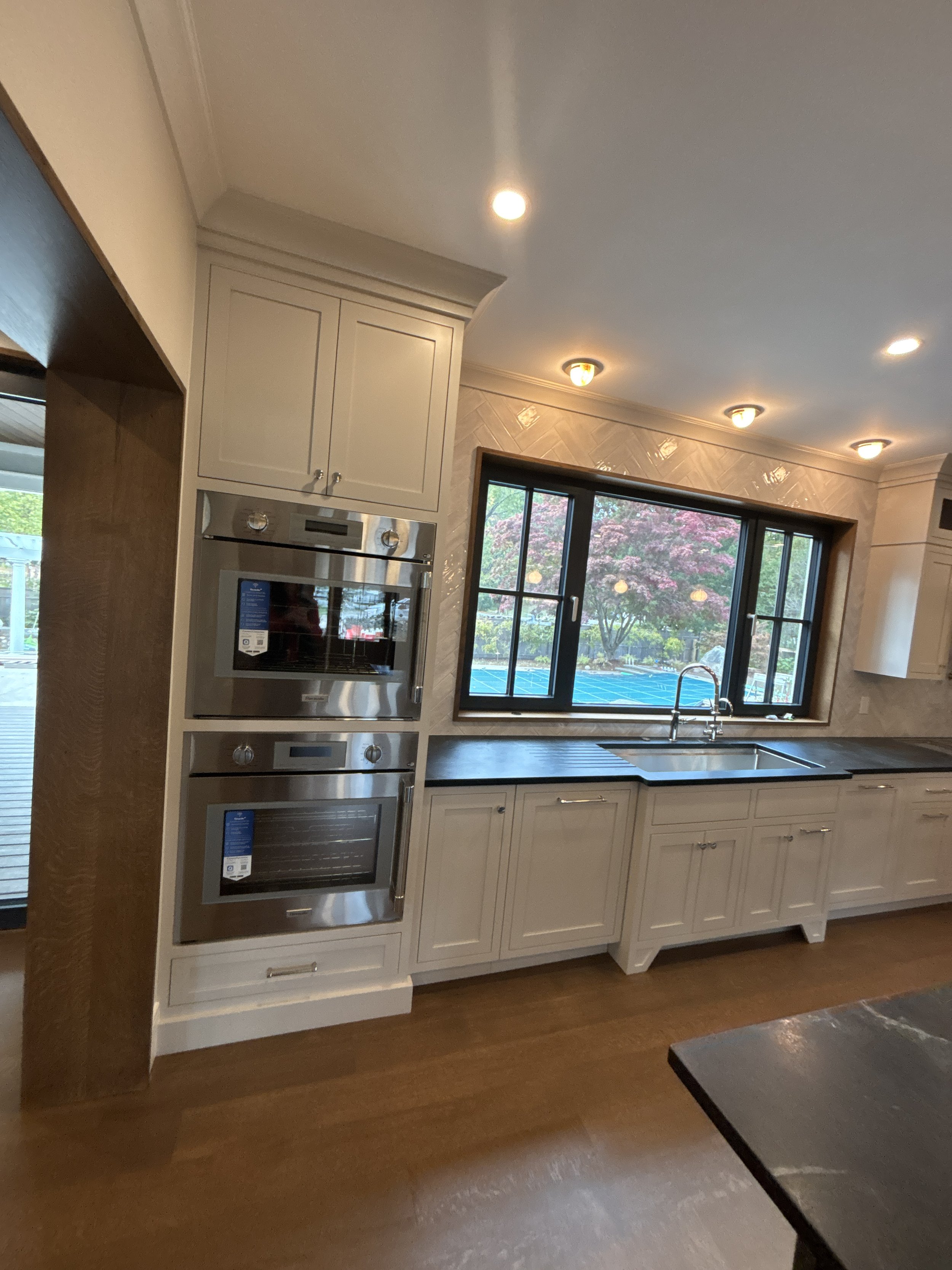 Kitchen with white cabinetry, stainless steel double ovens, a large Best general contractor, home builder, and home remodeler on the Northshore in Ando, and a view of a backyard with trees and a blue patio. Black countertops and a faucet are visible.