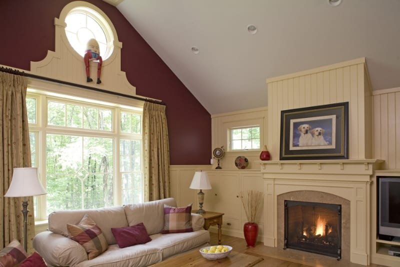 Living room with a cream-colored sofa, multiple pillows, large windows with beige curtains, a fireplace with a fire burning, framed dog's picture above the fireplace, small decorative items, lamps, and a red vase. A doll is placed on a small ledge ab