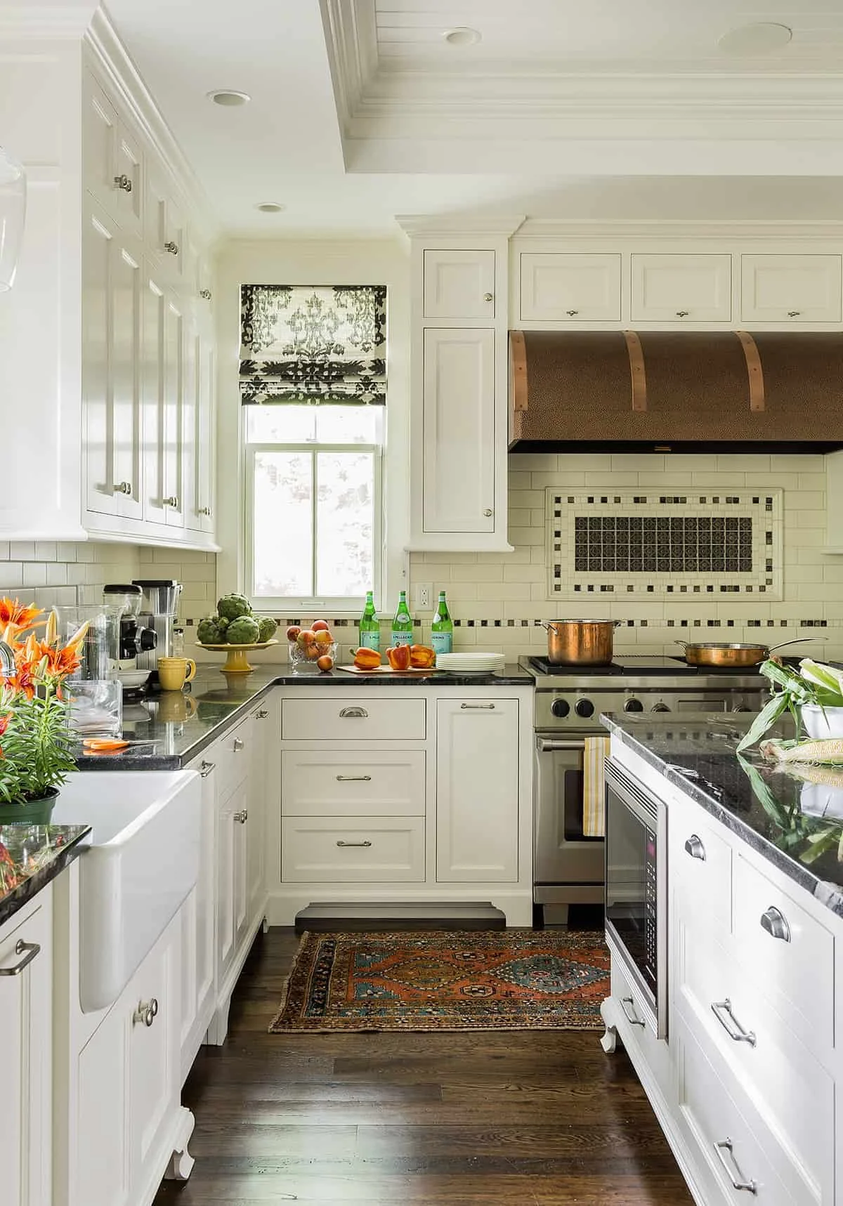 A bright, white kitchen with dark wood flooring, white cabinets, and a window with black and white patterned curtains. The countertop has vegetables, drinks, and kitchen appliances. There is a rug on the floor and a gas stove with pots and a range ho