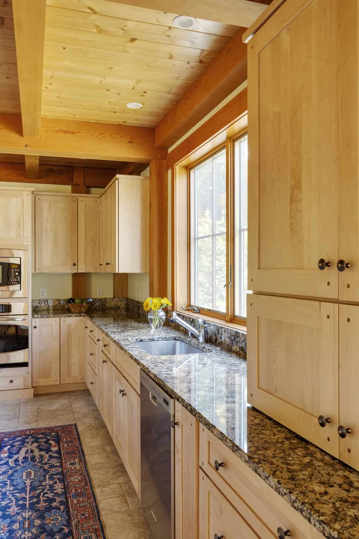 Kitchen with wooden cabinets, granite countertops, a window with natural light, a vase of yellow flowers on the counter, and a patterned rug on the floor.