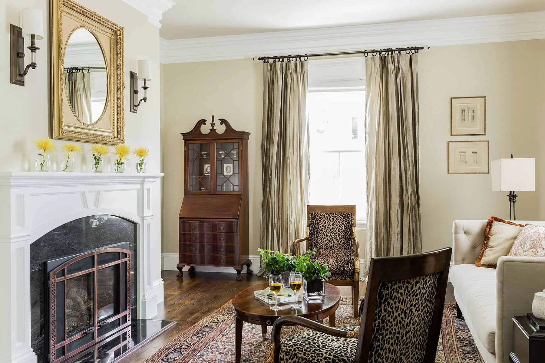Living room with white fireplace, gold framed mirror, wall sconces, beige walls, and a window with gold curtains. Furniture includes a leopard-print armchair, a white sofa with decorative pillows, wooden chairs, a small wooden table with two glasses 