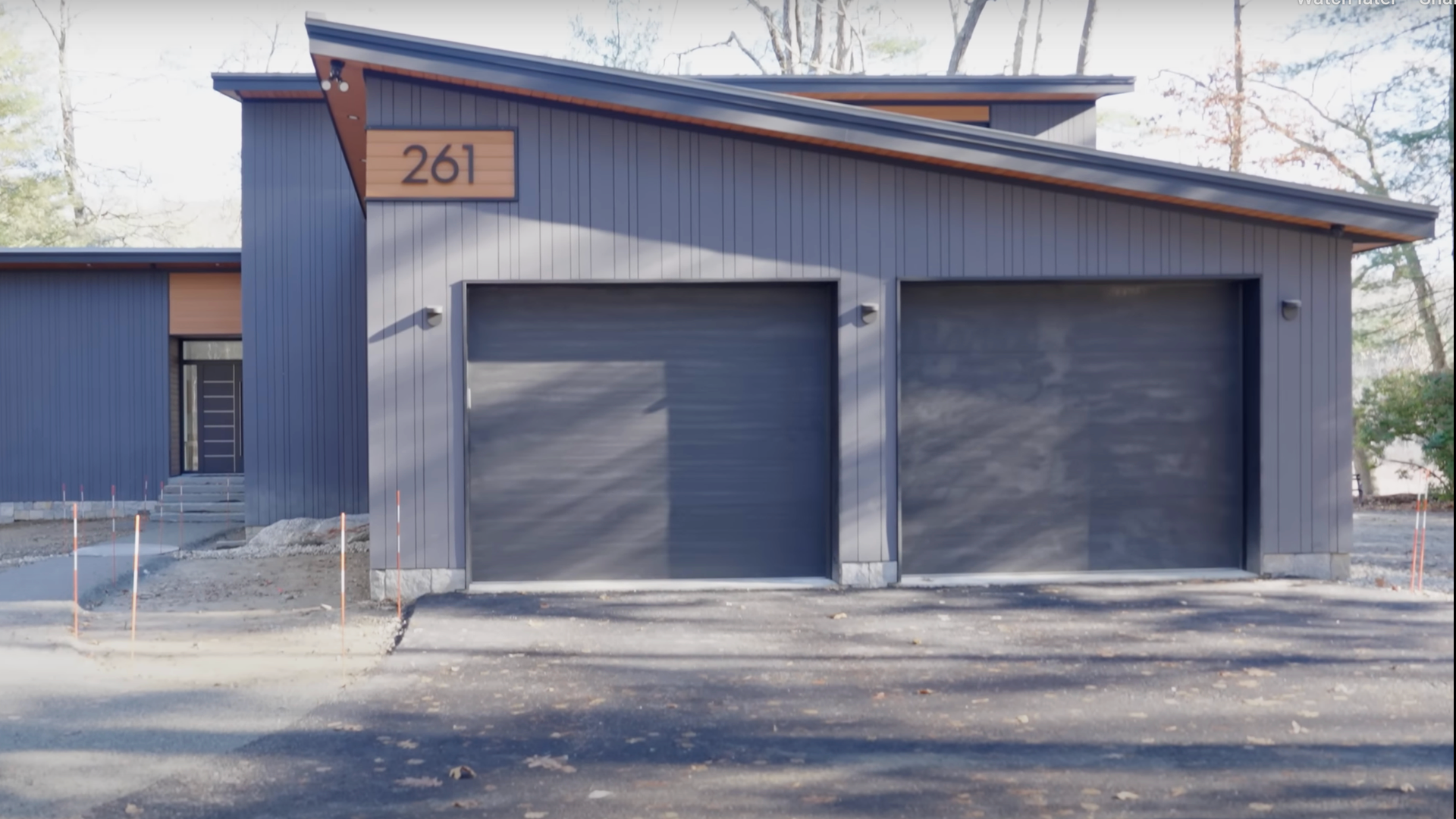 Front view of a modern house with two garage doors, blue vertical siding, featuring a wooden sign with the number 261, in a suburban area during daytime.