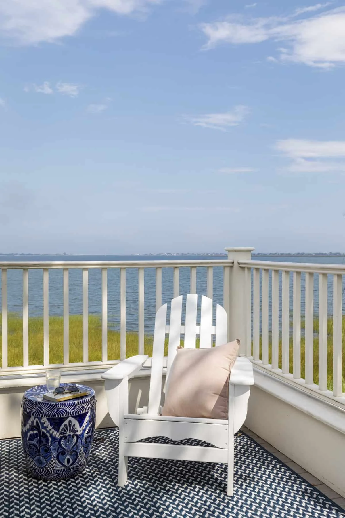 A white Adirondack chair with a pink pillow and a blanket on a balcony overlooking a body of water, with a railing and a cloudy sky in the background.