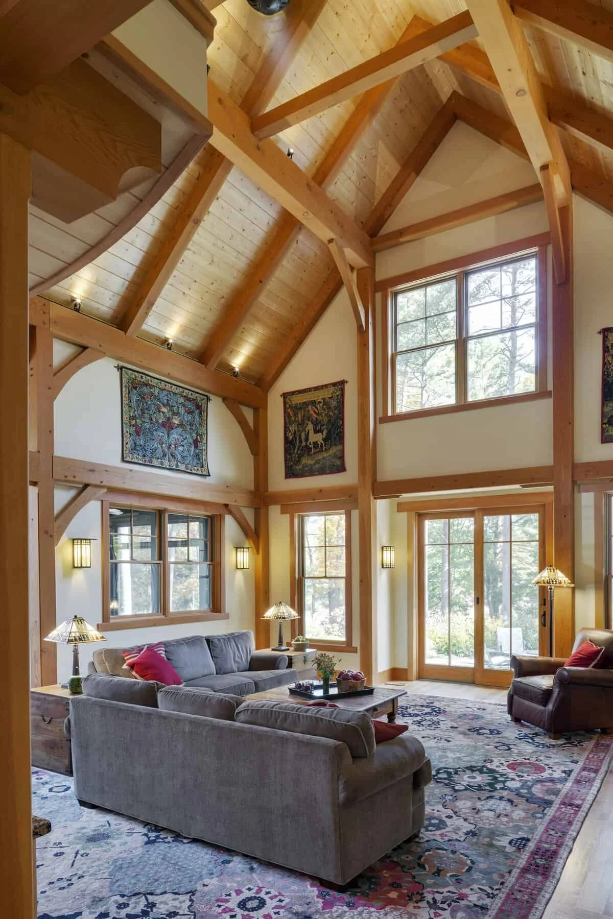 Living room with high vaulted ceiling, exposed wooden beams, large windows, and a colorful patterned area rug. Furniture includes a gray sofa, armchair, and coffee table, with wall art and floor lamps.