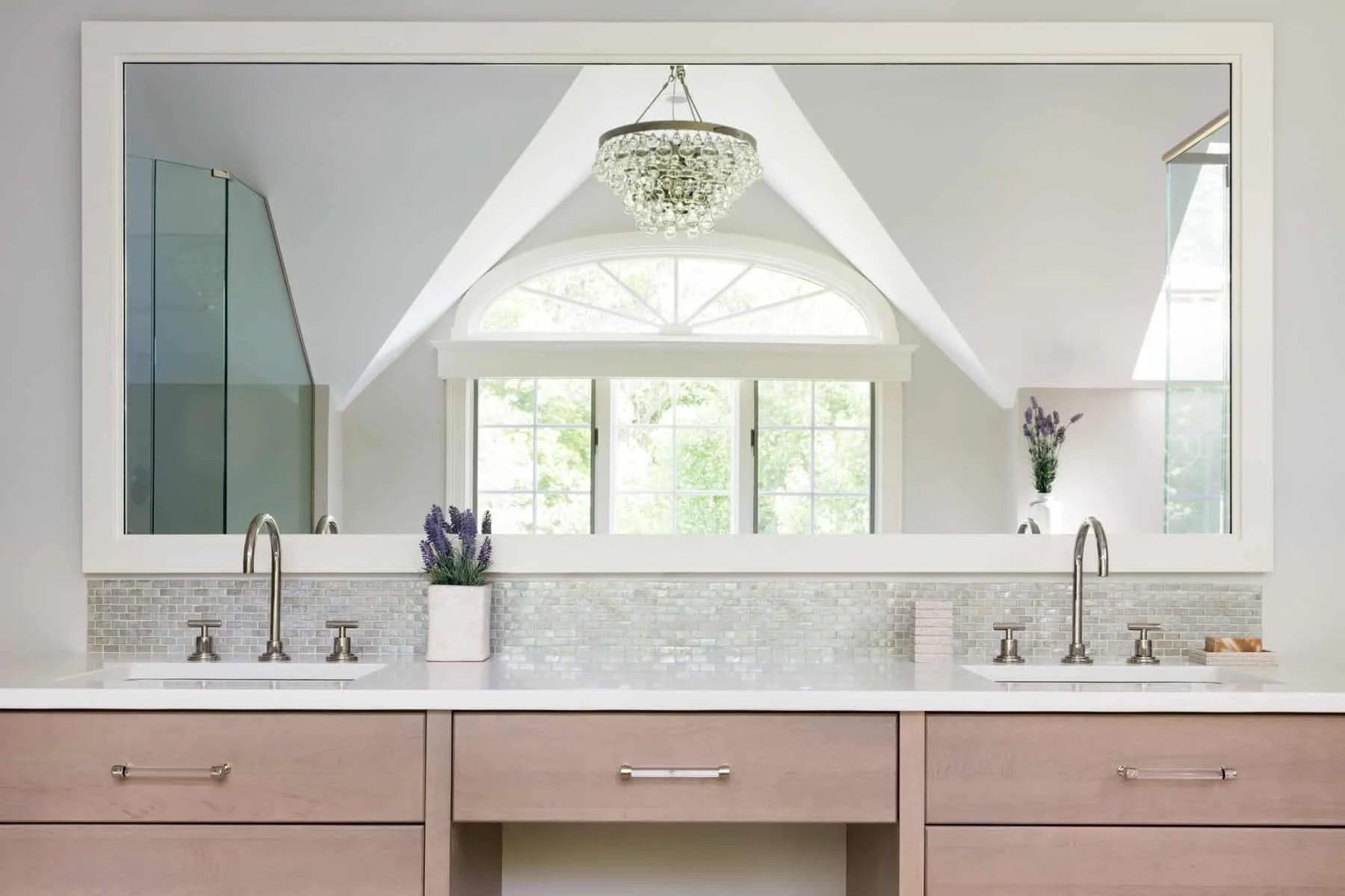 A bathroom vanity with a large mirror, two sinks, and a window reflecting natural light, decorated with lavender flowers.