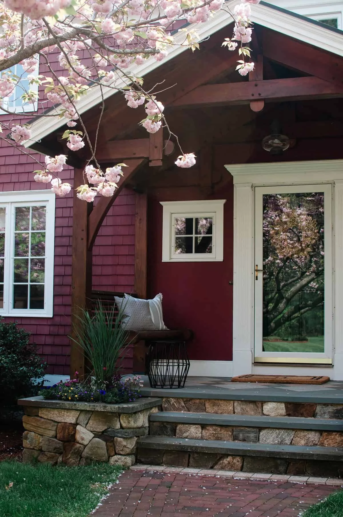 A porch with stone steps, a red house with white trim, a small window, and a glass door reflecting cherry blossom trees, with pink cherry blossoms in the foreground and hanging pot plants on the porch.