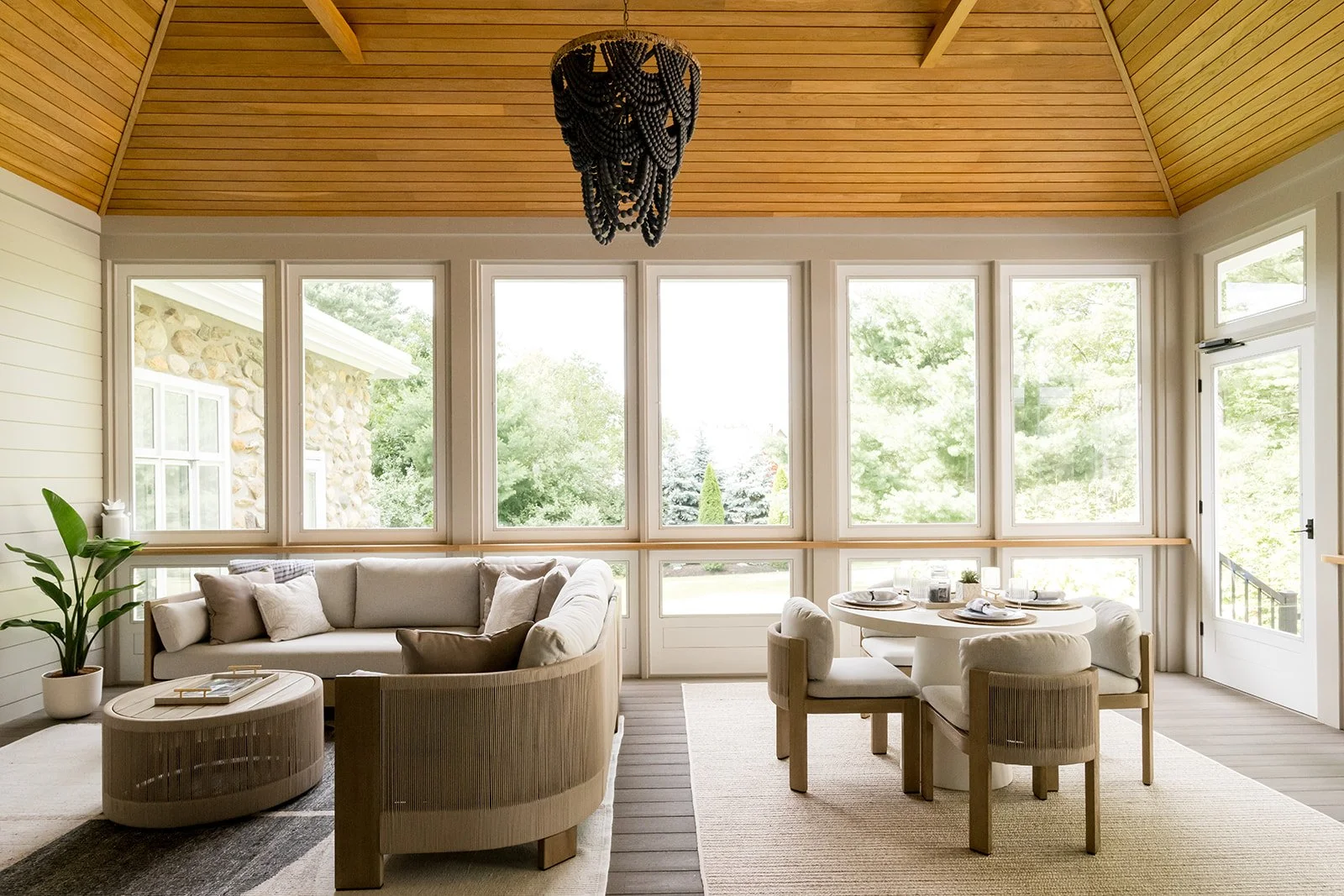 Bright living room with large windows, beige sofa with Cushions, round coffee table, round dining table with chairs, potted plant, and view of greenery outside.