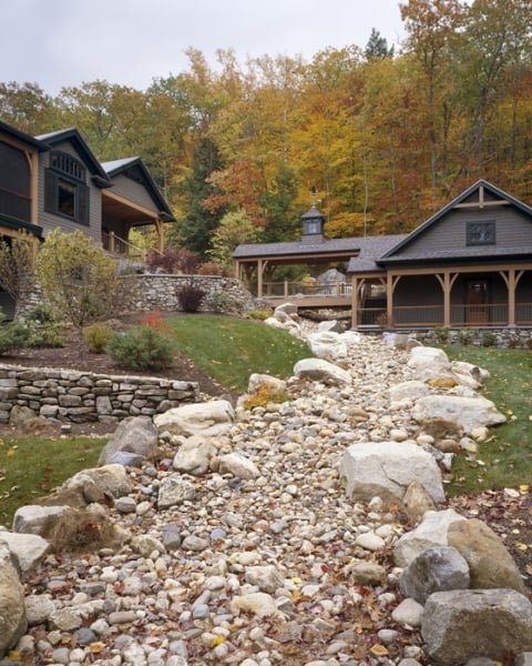 A dry creek bed with large rocks and pebbles runs through a landscaped yard with plants and bushes, leading to a house and a covered porch surrounded by fall-colored trees.
