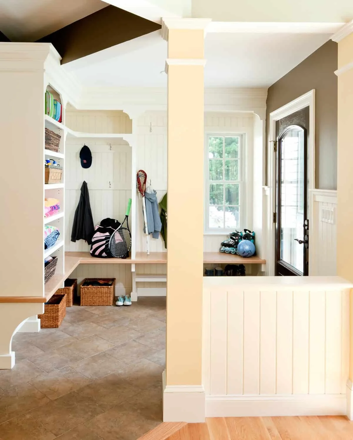 Foyer and mudroom area with coat hooks, baskets, and shoes. Natural light from a window, with a door leading outside, and sports equipment like rollerblades, a ball, and a helmet.
