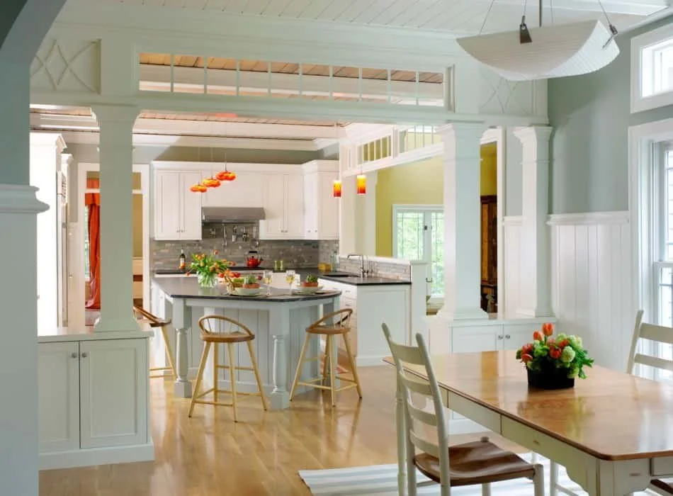 Bright kitchen with white cabinets, gray backsplash, and a central island with bar stools, adjacent to a dining area with a table and flower centerpiece.