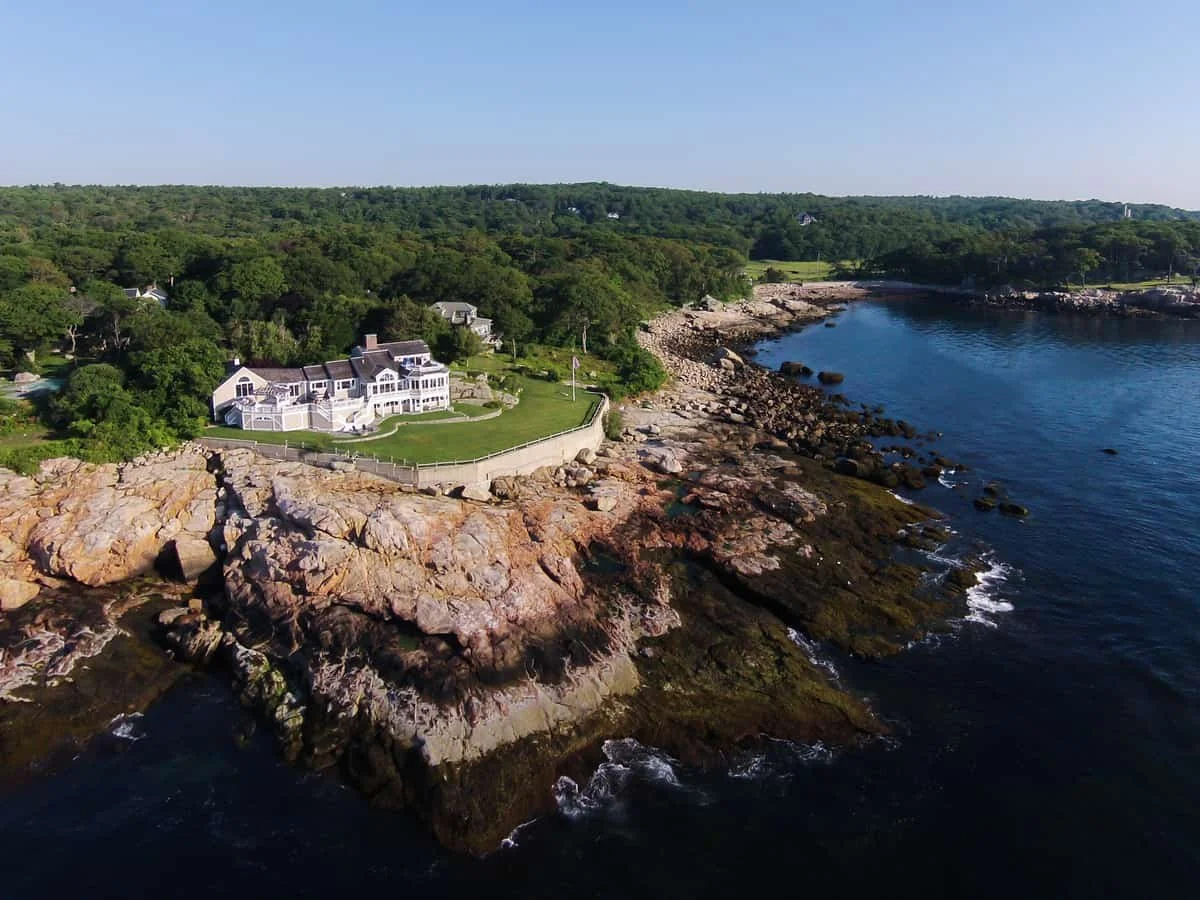 Aerial view of a coastal property with a large white house, green lawn, rocky shoreline, and calm ocean waters.