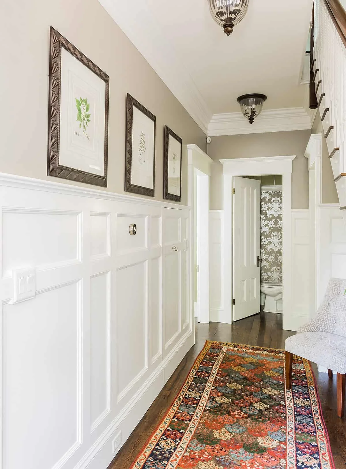 A hallway with white wainscoting, beige walls, and framed botanical art. There's a colorful oriental runner rug on hardwood flooring. A chair with light patterned upholstery is partially visible on the right. In the background, an open door reveals a