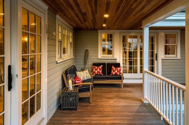 Covered outdoor porch with wooden flooring, white railings, and gray siding walls. Features a small seating area with black cushions, red and white pillows, a side table with candles, and a sliding glass door leading indoors.